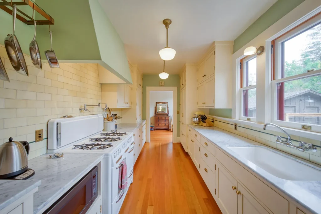 1111+Emerson+Street-13-1920w | Michael Meyer Construction + Fine Wood Working | Michael Meyer Construction + Fine Wood Working Bright, narrow kitchen with white cabinets, marble countertops, gas stove, hanging utensils, wood floor, large farmhouse sink, and three windows letting in natural light.