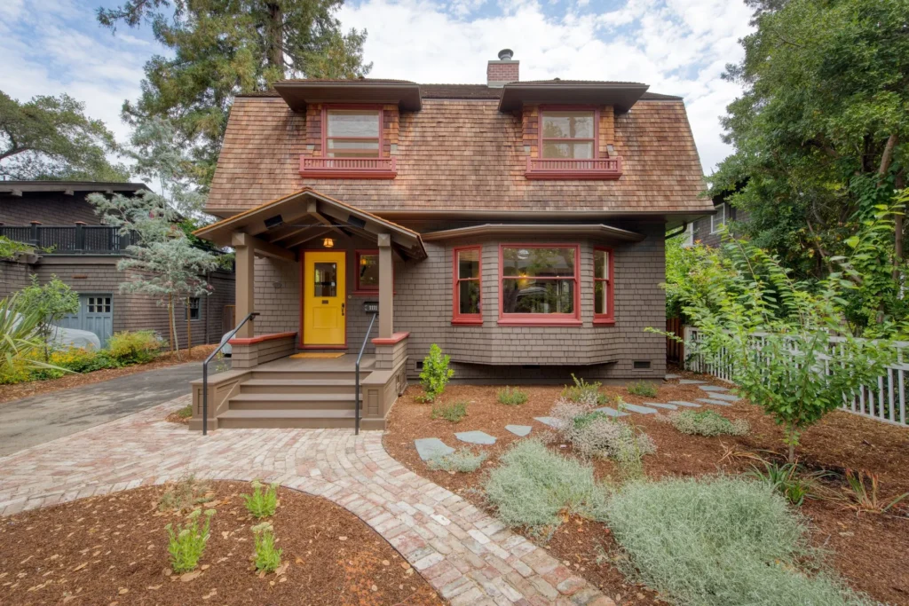 A two-story brown house with red trim, a yellow front door, a small porch, and a brick pathway, surrounded by plants and trees in the yard.