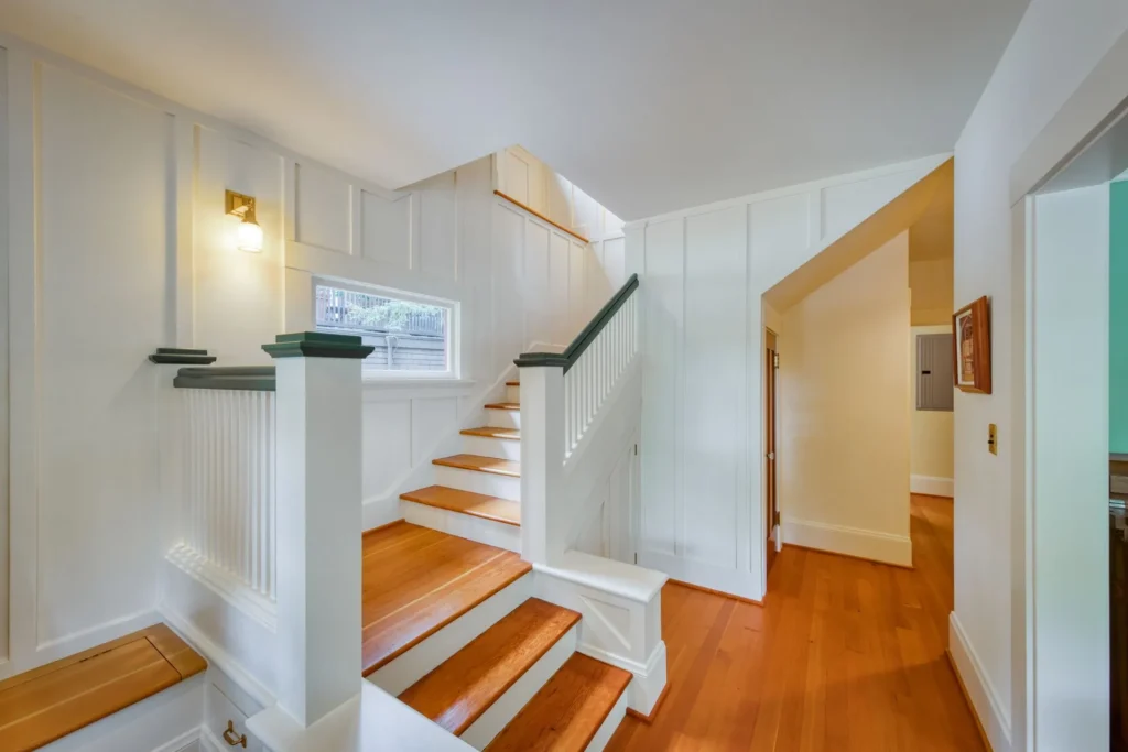 1111+Emerson+Street-6-1920w | Michael Meyer Construction + Fine Wood Working | Michael Meyer Construction + Fine Wood Working Bright wooden staircase with white paneled walls, green railings, and a small window. Warm wood floors extend into a hallway with angled ceilings, creating an inviting, well-lit interior.