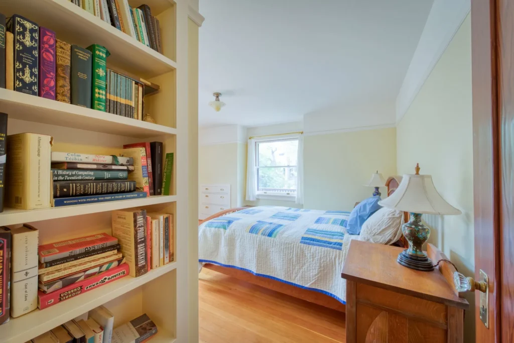 1111+Emerson+Street-8-1920w | Michael Meyer Construction + Fine Wood Working | Michael Meyer Construction + Fine Wood Working A cozy bedroom with a wooden floor, a bed with a blue and white quilt, a lamp on a bedside table, a window with daylight, and a built-in bookshelf filled with books in the foreground.