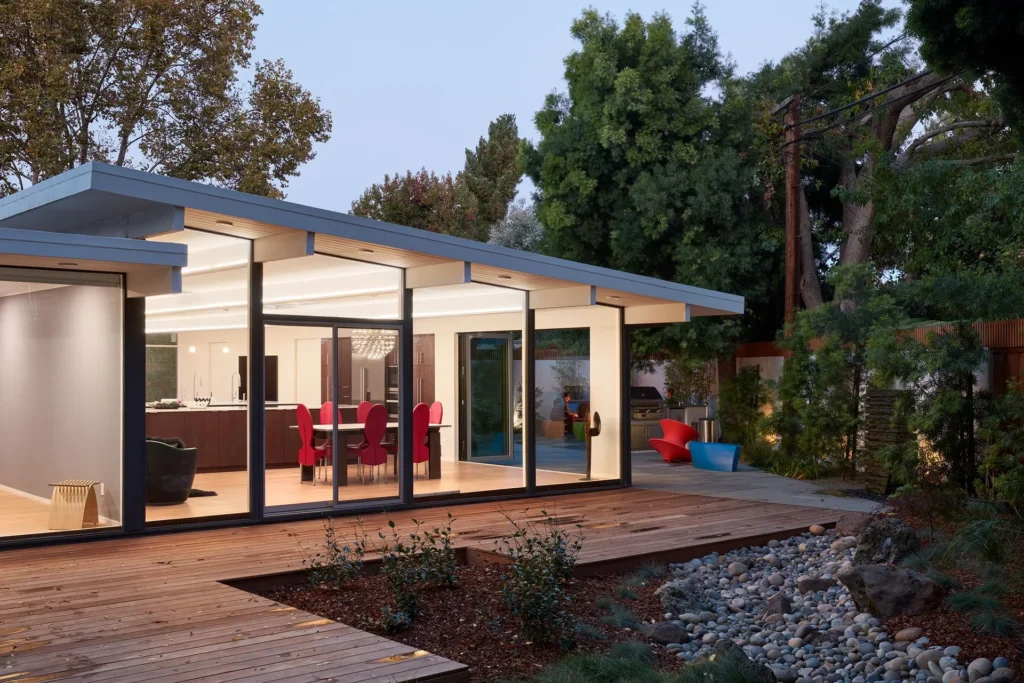 A modern house with large glass windows, an open dining area with red chairs, a wooden deck, and a landscaped yard with rocks and trees in the background.