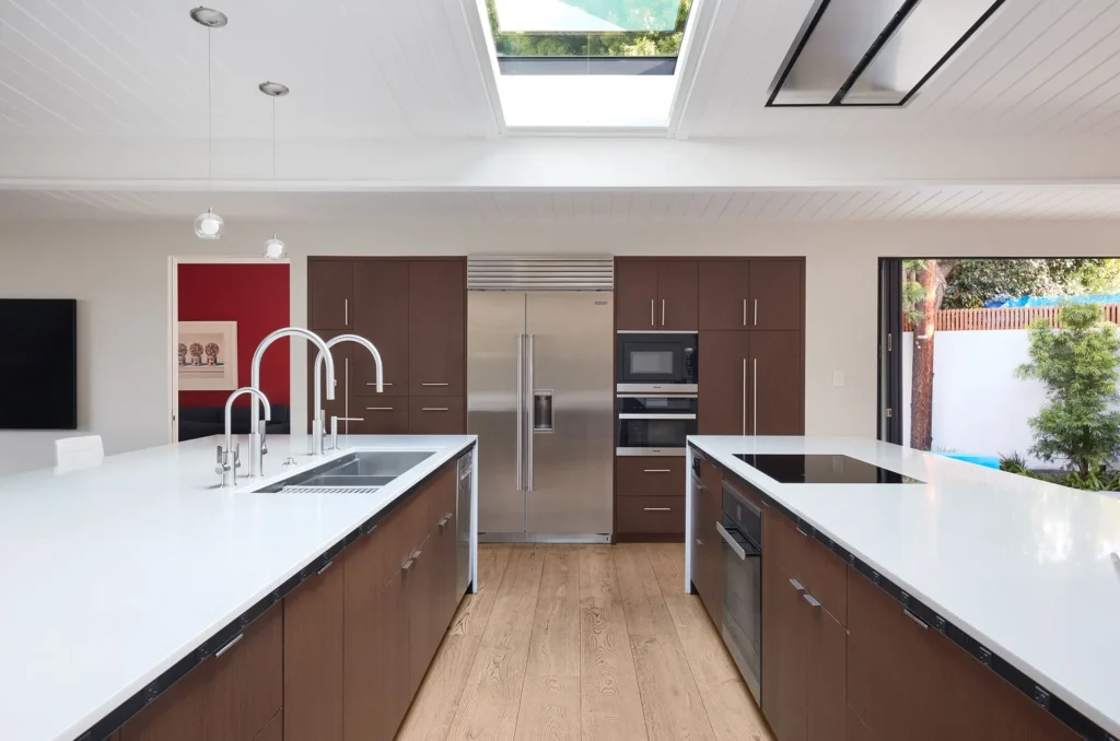 Modern kitchen with brown cabinets, white countertops, two islands, built-in stainless steel appliances, wood floor, and a skylight above letting in natural light.