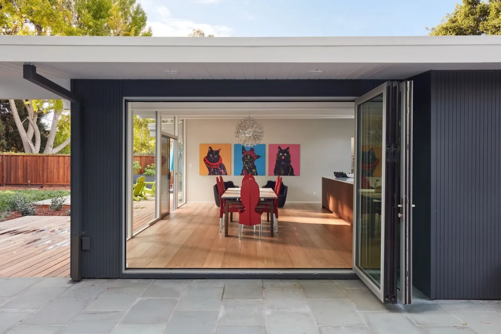 Modern dining room with red chairs, colorful cat art on the wall, large glass doors open to a patio, and wooden floors. Trees and a wooden fence are visible outside. Contemporary decor and bright, natural light fill the space.