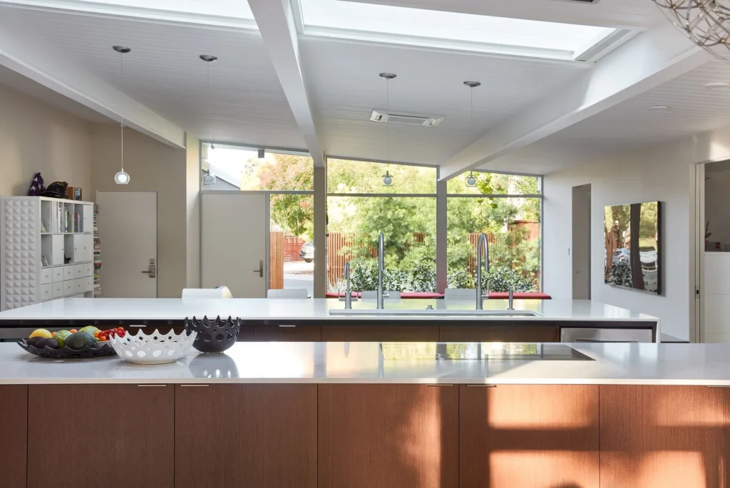 Modern kitchen with wood cabinets, a large white island, fruit bowls, pendant lights, skylights, and floor-to-ceiling windows revealing trees and a patio outside. Artwork hangs on the wall to the right.