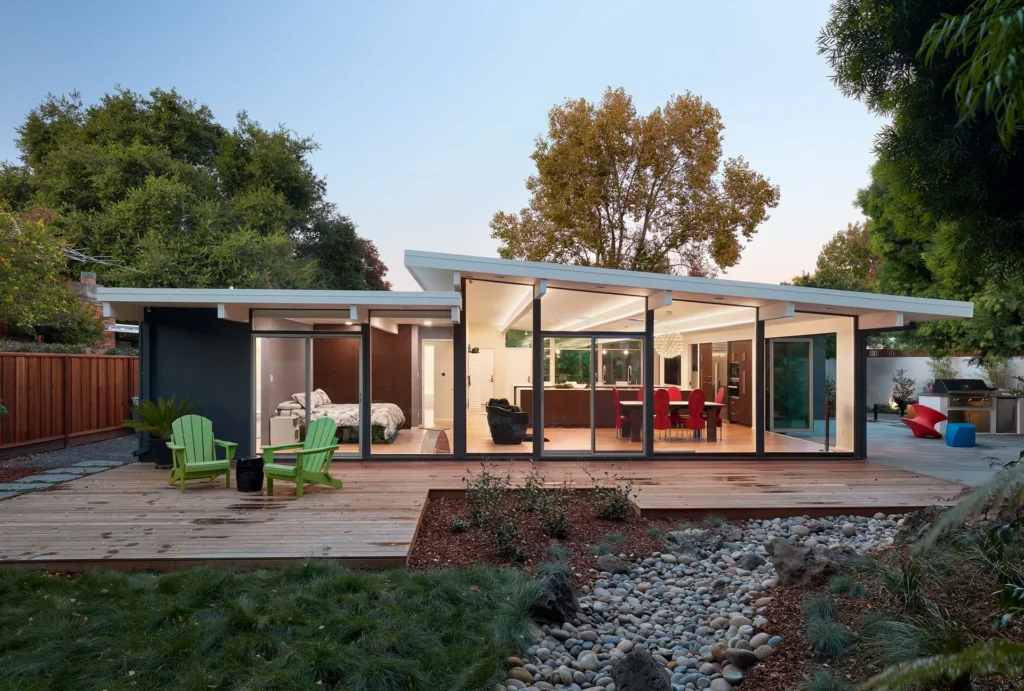 Modern one-story house with large glass windows, wooden deck, two green chairs, and landscaped yard with rocks and grass. The interior is brightly lit, showing a dining area and bedroom. Trees surround the home.