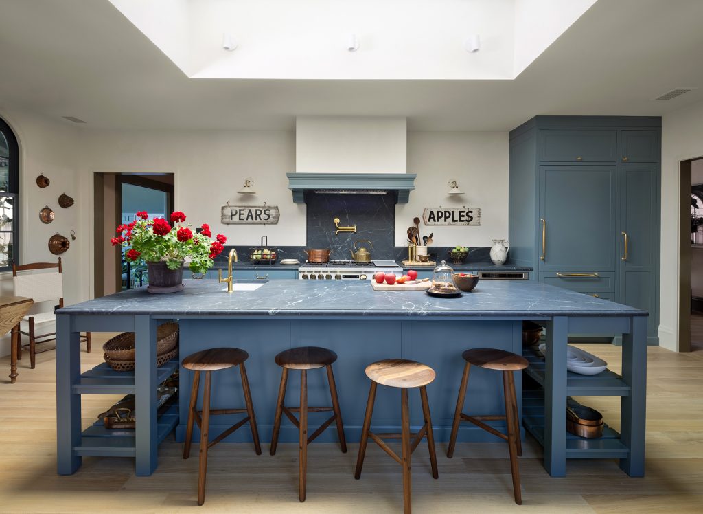 A modern kitchen with blue cabinets, a marble island, four wooden stools, a vase of red flowers, and signs reading PEARS and APPLES above a stove with gold fixtures. Natural light fills the space.