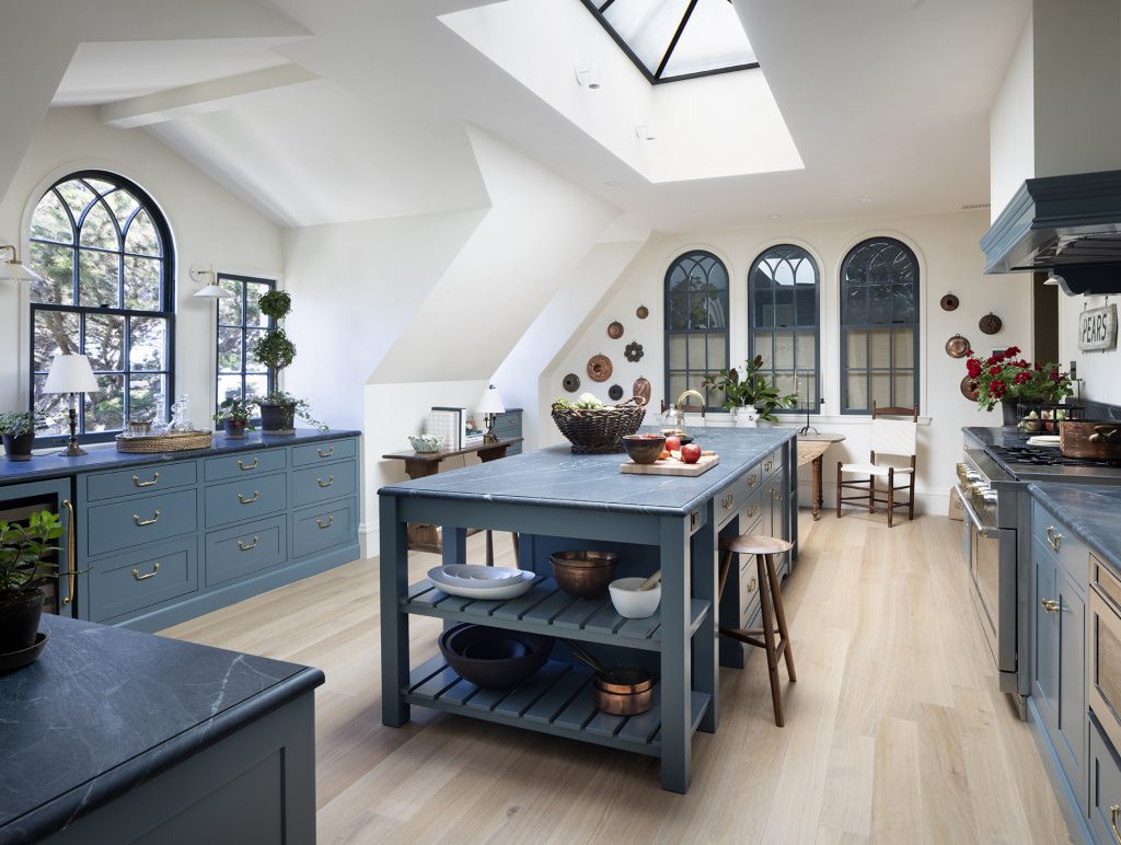 Bright kitchen with blue cabinets, marble countertops, large island, skylights, arched windows, wood floors, open shelves, and potted plants. Fruits and kitchenware decorate the counters, creating a cozy and elegant atmosphere.