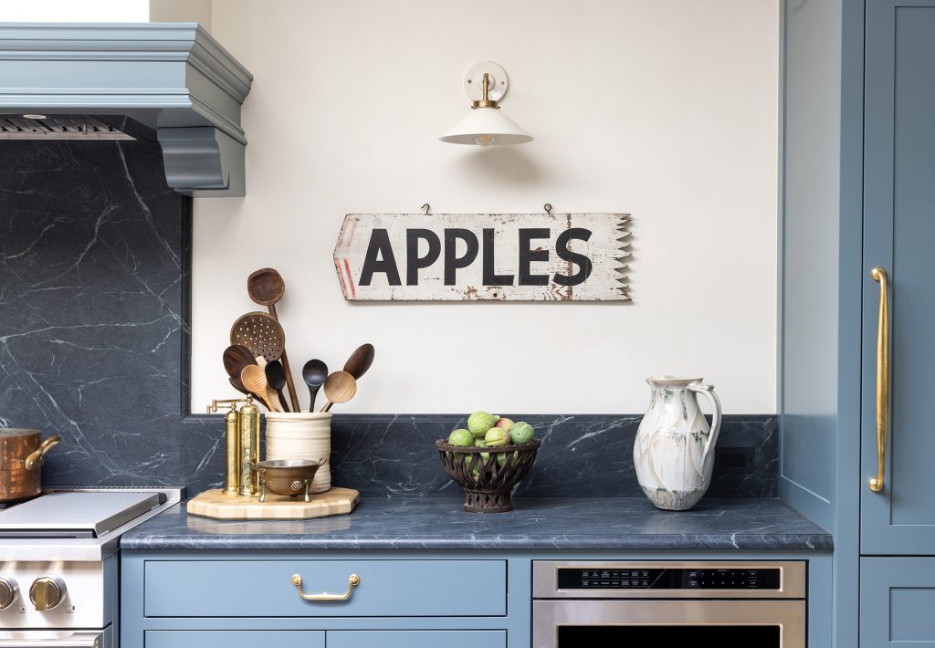 A kitchen with blue cabinets, a marble countertop, utensils in a holder, a bowl of green apples, a ceramic jug, and a rustic sign reading APPLES on the wall.