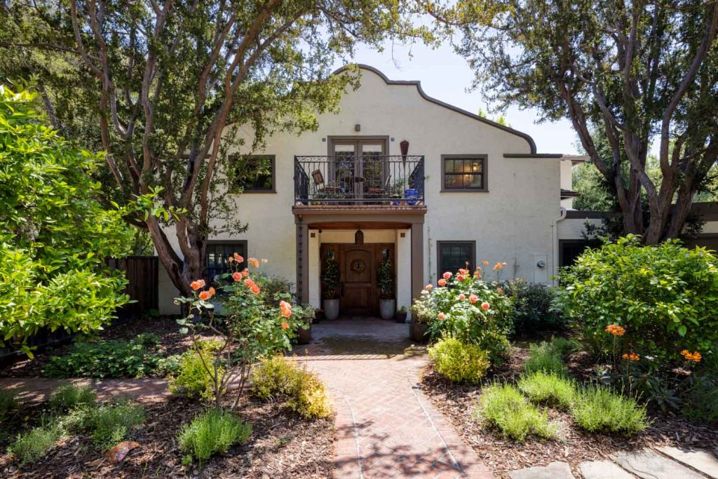 Creekside-1 | Michael Meyer Construction + Fine Wood Working | Michael Meyer Construction + Fine Wood Working Two-story stucco house with a balcony, arched roofline, and a wooden front door, surrounded by lush gardens, blooming flowers, brick walkway, and tall trees.