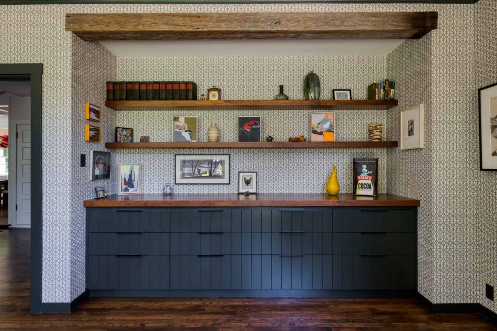 Creekside-10 | Michael Meyer Construction + Fine Wood Working | Michael Meyer Construction + Fine Wood Working A built-in dark green cabinet with a wood countertop sits below two wooden shelves displaying books, framed photos, decor, and a yellow vase, against patterned wallpaper.