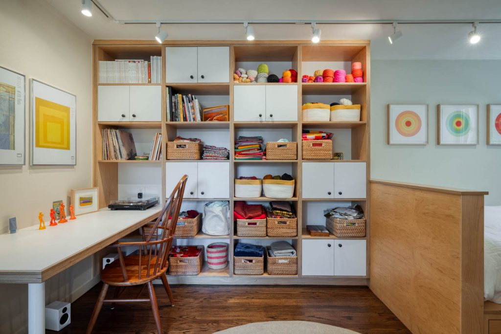 Organized craft room with a wooden desk and chair, open shelving holding books, yarn, fabric, baskets, and art supplies. Framed artwork hangs on the walls; wood floors and track lighting brighten the space.