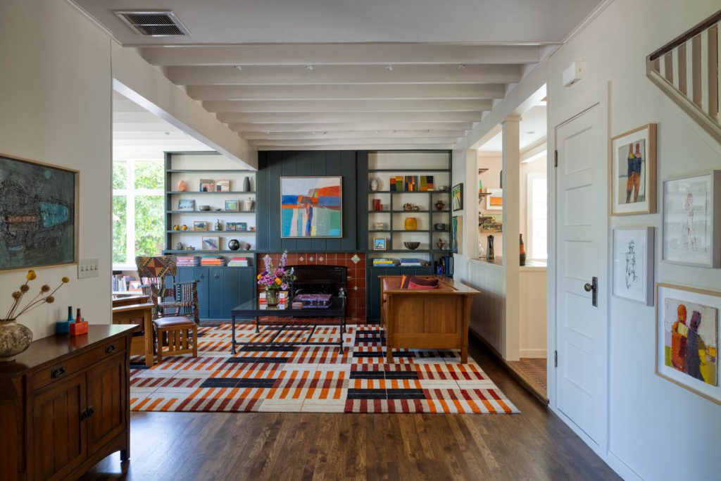Creekside-4 | Michael Meyer Construction + Fine Wood Working | Michael Meyer Construction + Fine Wood Working Bright, colorful living room with patterned rug, modern artwork, wooden furniture, a fireplace, open shelves with decor, and large windows letting in natural light. Walls and ceiling are white with wood accents.