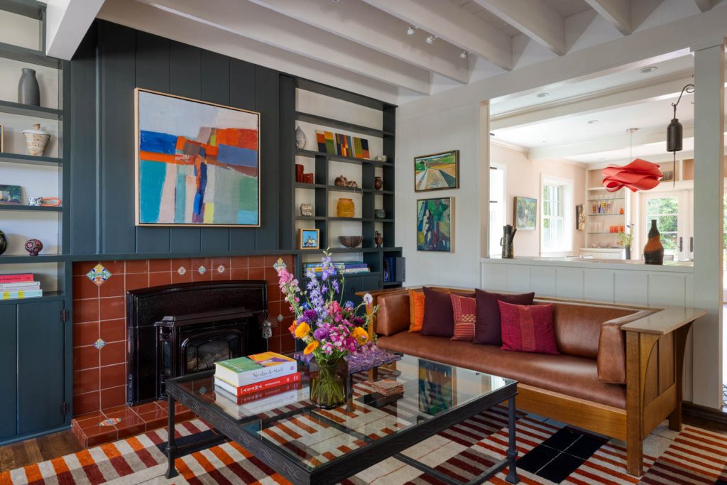Cozy living room with a brown leather sofa, colorful pillows, glass coffee table with books and flowers, geometric rug, shelves with decor, modern abstract art, and a fireplace. Open to a bright adjacent room.