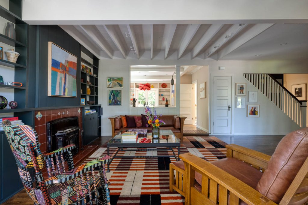 Creekside-7 | Michael Meyer Construction + Fine Wood Working | Michael Meyer Construction + Fine Wood Working Bright living room with colorful patterned rug, armchairs, a glass coffee table with flowers, built-in shelves, a fireplace, modern art, and exposed ceiling beams. Stairs and hallway visible in the background.