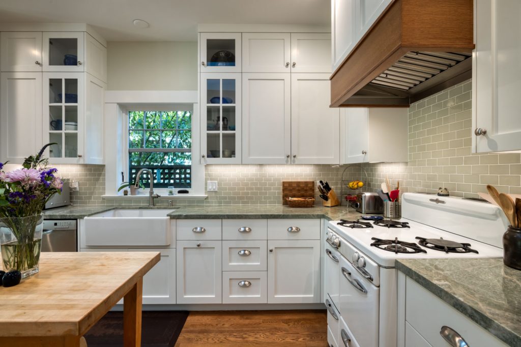 Bright kitchen with white cabinets, green countertops, farmhouse sink, gas stove, and wood accents. A window above the sink lets in natural light. Flowers and utensils decorate the counters.