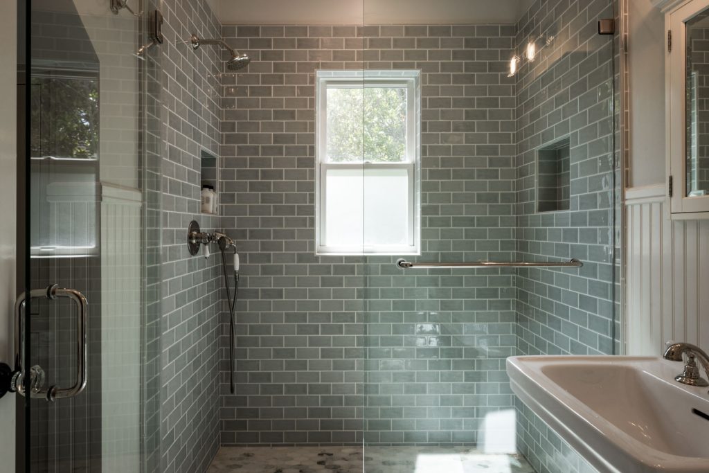 A modern bathroom shower with greenish-gray subway tiles, a glass door, a window letting in light, metal fixtures, a built-in shelf, and a white rectangular sink to the right.