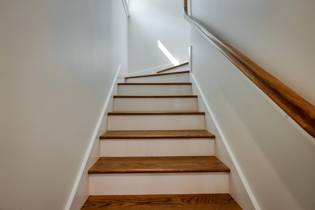 Wooden stairs with white risers and a brown handrail on the right, leading upward in a bright, narrow hallway. Natural light enters from a window at the top of the stairs.