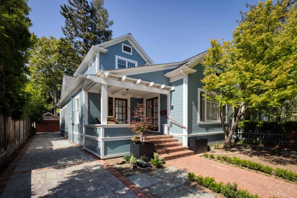 A blue house with white trim, a covered front porch, red brick steps, driveway on the left, trees and shrubs around, and a detached garage in the background on a sunny day.