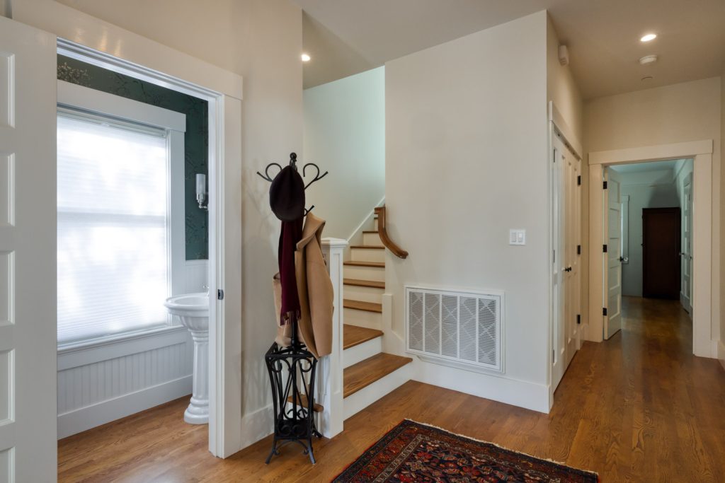 A hallway with wooden floors, a staircase, a coat rack holding a hat, scarf, and coat, and a bathroom with a pedestal sink visible through an open door. Natural light comes through a frosted window.